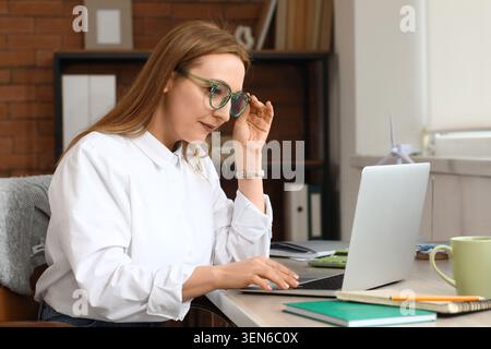 Donna concentrata che regola gli occhiali da vista mentre lavora su un notebook in un moderno ambiente di ufficio domestico Foto Stock