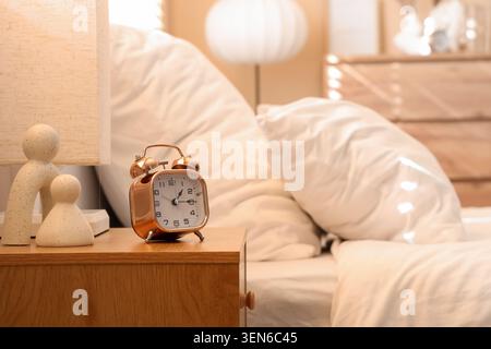 Sveglia in rame su comodino in legno con letto e lampada, sveglia mattutina, interno della camera da letto Foto Stock