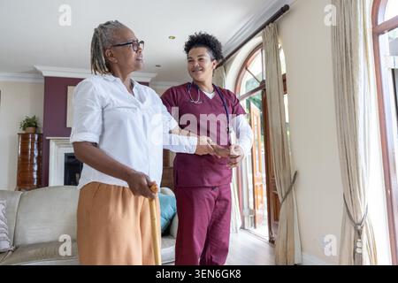 Badante negli scrub di colore marrone che aiuta una donna afroamericana anziana a fare il passo a casa tenendo il bastone Foto Stock