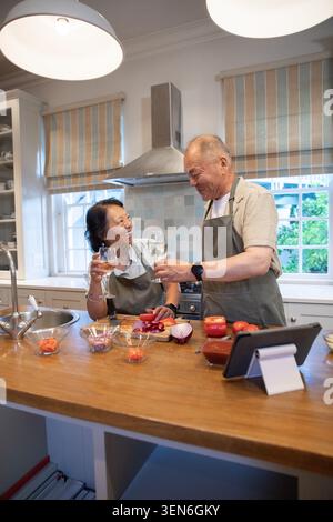 Donna asiatica anziana e uomo che indossano grembiuli grigi a Kitchen Island dopo tavoletta e tostatura di vino Foto Stock