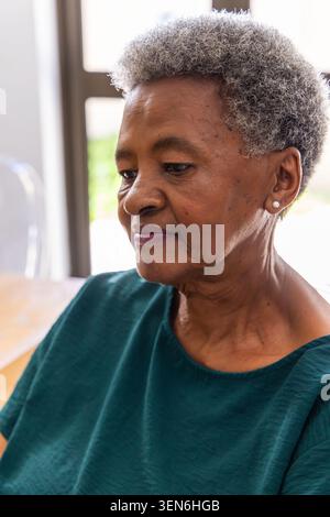 Donna afroamericana anziana seduta al tavolo vicino alla porta di vetro, guardando verso il basso, indossando una teal top Foto Stock