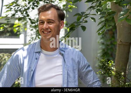 Uomo adulto in piedi sorridente nel cortile sotto la luce del sole appannata con tee bianche e camicia a righe Foto Stock