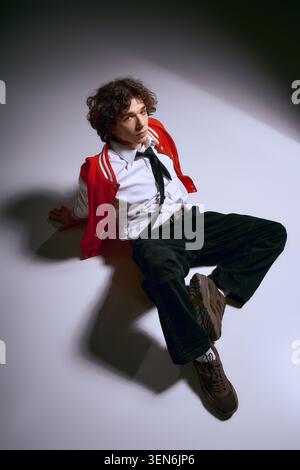 Bel giovane con i capelli ricci si siede graziosamente sul pavimento, mostrando il suo stile alla moda. Foto Stock
