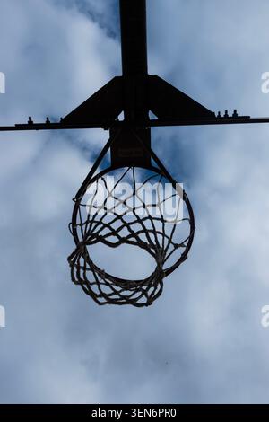 Vista ad angolo basso di un canestro da basket all'aperto e silhouette a rete su un cielo blu nuvoloso Foto Stock
