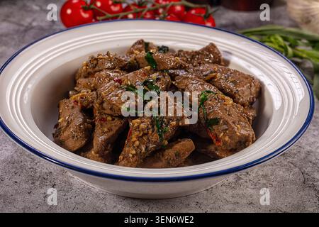 Fette di fegato di manzo cotte saporite condite con spezie alle erbe, servite in un recipiente di ceramica bianca con bordo blu. Pomodori freschi cipolle verdi sullo sfondo Foto Stock
