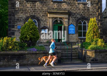 Persona (vista laterale) e cane nel centro del villaggio di Addingham (dopo l'attraente edificio tradizionale in pietra soleggiata) - Main Street, West Yorkshire, Inghilterra, Regno Unito. Foto Stock