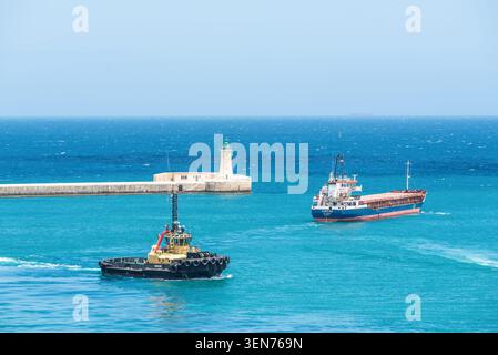 La Valletta, Malta - 16 maggio 2025: Nave da carico e rimorchiatore in barca a vela vicino allo storico ingresso del porto e al faro di la Valletta, Malta, circondato da bricco Foto Stock
