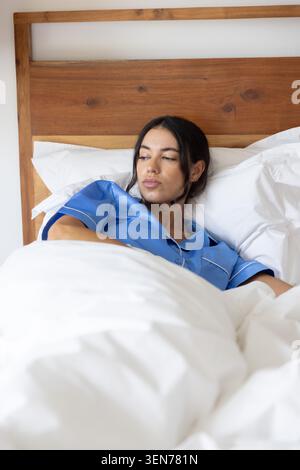 Donna bianca che indossa pigiami blu sdraiati in camera da letto contro testiera in legno cuscini bianchi, piumone Foto Stock