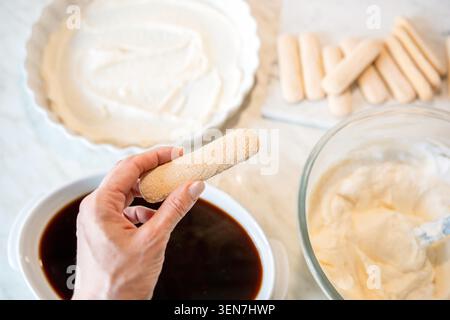 Donna che immerge a mano il biscotto al ladyfinger nel caffè mentre prepara il tiramisù. Processo di dessert fatto in casa con panna e ingredienti sulla superficie della cucina, spazio per copiare Foto Stock