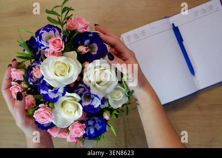 Le mani femminili tengono in mano un fiore di rosa. Quaderno, diario con penna su un tavolo di legno vicino a un mazzo di rose multicolore. Foto Stock