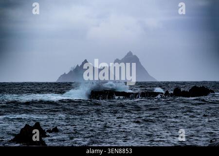 La lontana isola di Skellig Michael è avvolta dalla nebbia, vista dall'aspra costa irlandese con onde che si infrangono e rocce taglienti in primo piano Foto Stock
