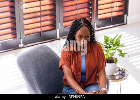 Donna africana seduta su una poltrona grigia in un soggiorno illuminato dal sole, indossa camicia ruggine, braccialetti, guarda Foto Stock