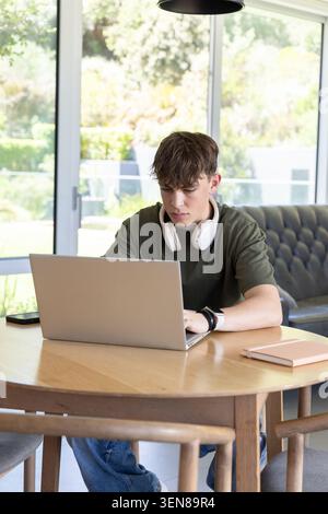 Un maschio adolescente seduto a un tavolo rotondo di legno che lavora su un computer portatile con cuffie e smartphone Foto Stock