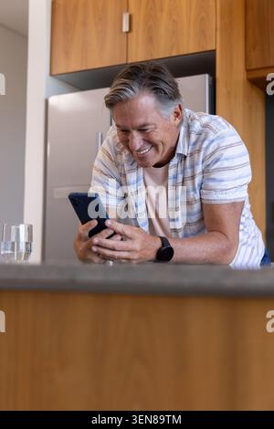 Un uomo anziano appoggiato in avanti sul bancone della cucina, sorridendo e controllando lo smartphone con l'orologio da polso Foto Stock