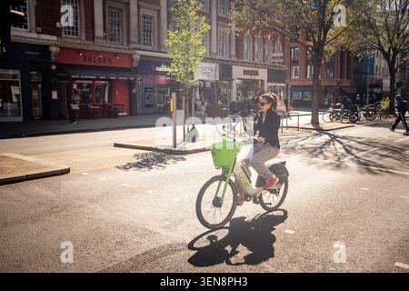 LONDRA - 22 APRILE 2026: HIGH STREET KENSINGTON - giovane donna che guida una bicicletta elettrica verde Lime sulla soleggiata strada di Londra Foto Stock
