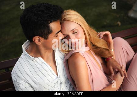 Una coppia amorevole si rilassa su una panchina, condividendo sorrisi e abbracciando gentilmente sotto il sole caldo. Foto Stock