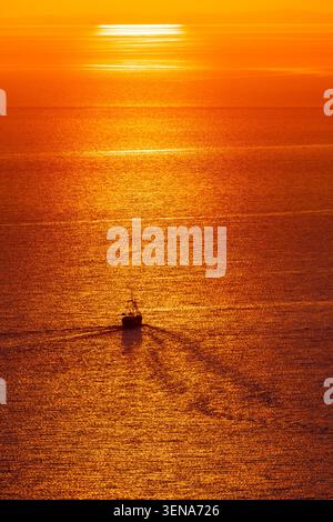Piccola nave che attraversa il mare dorato al tramonto con un lungo riflesso e la luce arancione del sole Foto Stock