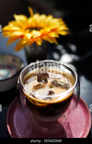 Caffè con ghiaccio in vetro trasparente sul tavolo con fiori nelle vicinanze in un caffè durante il giorno di sole Foto Stock