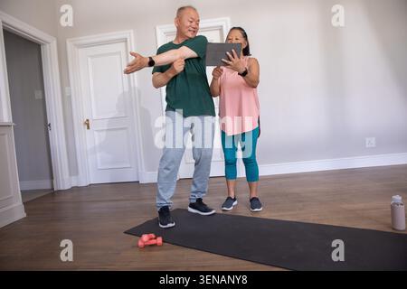 Donna cinese anziana e uomo cinese che si allunga in abbigliamento sportivo utilizzando tablet su tappetino con pesi rosa Foto Stock