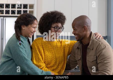 Famiglia diversificata seduta al bancone della cucina, madre con maglione in maglia gialla, uomo in giacca abbronzata sorridente Foto Stock