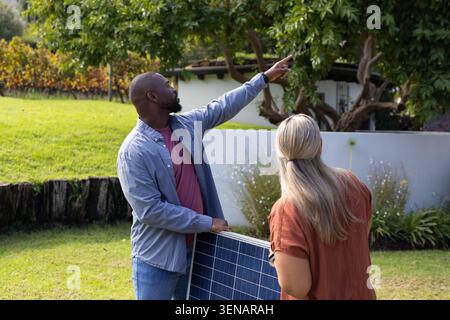 Uomo e donna di medie dimensioni con un grande pannello solare che punta verso il tetto di un prato residenziale Foto Stock