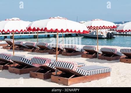 Una fila di eleganti sdraio in legno con cuscini a righe blu e bianche sotto ombrelloni bianchi abbinati con bordi orlati rossi su una spiaggia sabbiosa, con Foto Stock
