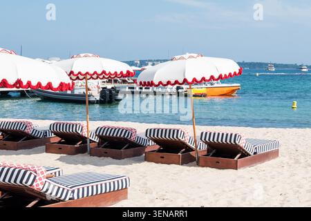 Una fila di eleganti sdraio in legno con cuscini a righe blu e bianche sotto ombrelloni bianchi abbinati con bordi orlati rossi su una spiaggia sabbiosa, con Foto Stock