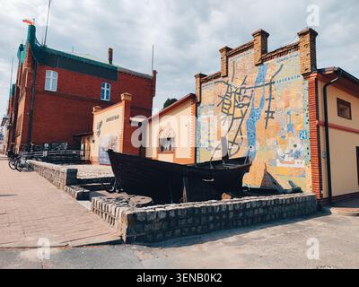 Wolin, Polonia, 13.08.2022: Barca storica in legno e mostra murale. Scena all'aperto con un edificio coperto di murales, una vecchia barca in legno in mostra, mattoni A. Foto Stock
