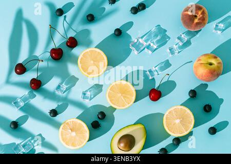 Scena estiva con fette di limone, ciliegie, mirtilli, pesche, avocado, cubetti di ghiaccio e ombra di foglie di palma su sfondo blu. Foto Stock