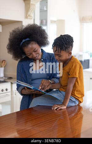 Madre e figlio afroamericani seduti al banco della cucina in legno lucido leggendo un libro illustrato Foto Stock