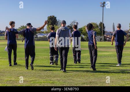 Squadra di cricket maschile che cammina attraverso il campo erboso indossando camicie navy con piazzole e appunti Foto Stock