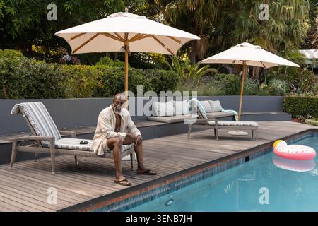 Uomo africano maturo che si rilassa a bordo piscina sul ponte di legno indossando camicia aperta, sandali e occhiali da sole nelle vicinanze Foto Stock
