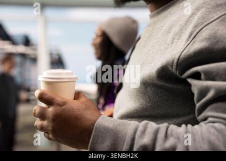 Primo piano del passeggero dell'aeroporto che tiene in mano una tazza di caffè da asporto accanto ai viaggiatori che camminano su sfondo sfocato. Contenitore per bevande in mano turistica che naviga nel centro di trasporto dell'aeroporto Foto Stock