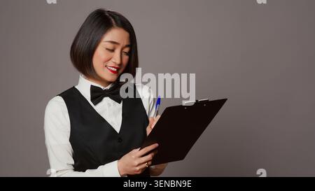 Concierge asiatica che crea una lista di controllo delle registrazioni, lavora su prenotazioni online e prenotazioni in studio, receptionist che guarda i documenti degli appunti su sfondo grigio. Foto Stock