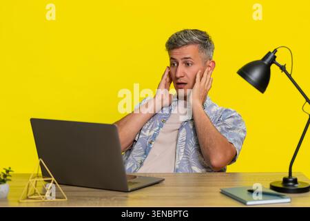 Uomo di mezza età che legge messaggi e-mail inaspettati sul laptop a tavola, mostrando attenzione, poi si piega improvvisamente con gli occhi spalancati. Giovane ragazzo che reagisce emotivamente per sorprendere su sfondo giallo. Foto Stock