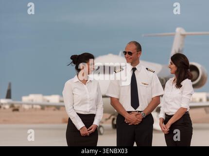 Cordiale equipaggio privato con pilota sorridente e due assistenti di volo in uniforme bianca che parlano sull'asfalto dell'aeroporto al crepuscolo dorato accanto a un'azienda Foto Stock