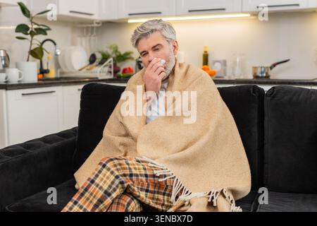 Uomo di mezza età a casa divano sotto tosse plaid salvietta il naso con brividi di tessuto e sniffles durante un'influenza dura, starnuti salviette non in tovagliolo sente freddo soffrendo di allergia sintomi influenzali quarantena. Foto Stock
