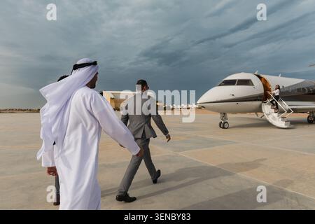 Uomo d'affari sicuro degli Emirati in kandura tradizionale e adatto a un dirigente che cammina verso un jet privato sull'asfalto dell'aeroporto sotto il cielo spettacolare, ideale per Foto Stock