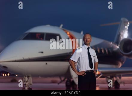 Pilota di linea maschile sicuro in piedi in uniforme con le mani sui fianchi sull'asfalto dell'aeroporto accanto a jet privati di lusso al tramonto, ideale per la carriera nell'aviazione e. Foto Stock
