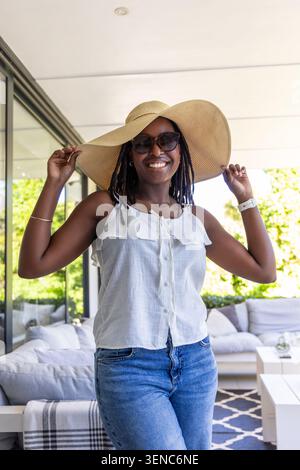 Donna afro-americana in piedi sul patio che regge il cappello, indossa gli occhiali da sole, dietro un divano esterno Foto Stock