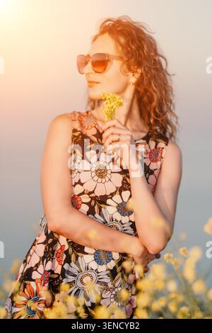 Donna fiori estate, bella giovane donna con capelli rossi ricci che tengono i fiori gialli in un campo all'aperto di un'ora d'oro al tramonto Foto Stock