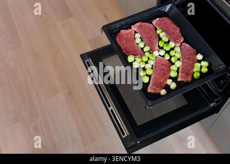 Bistecche di maiale crude e cavoletti di Bruxelles su vassoio del forno pronti per la tostatura in cucina, attenzione selettiva Foto Stock