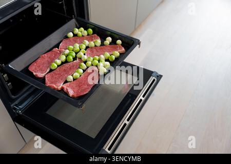 Bistecche di maiale crude e cavoletti di Bruxelles su vassoio del forno pronti per la tostatura in cucina, attenzione selettiva Foto Stock