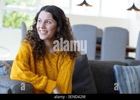 Donna seduta su un divano grigio vicino a una grande finestra, che indossa un maglione giallo senape, sorridente Foto Stock