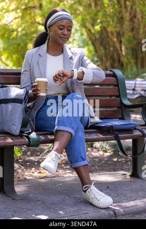 Donna seduta sulla panchina del parco che tiene la tazza, controlla smartwatch, zaino, borsa blu, sneakers bianche Foto Stock