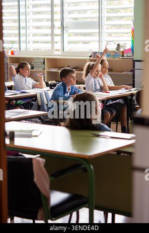 Gruppo di studenti delle scuole primarie che partecipano attivamente a una lezione in classe alzando le mani per rispondere alla domanda di un insegnante. Foto Stock