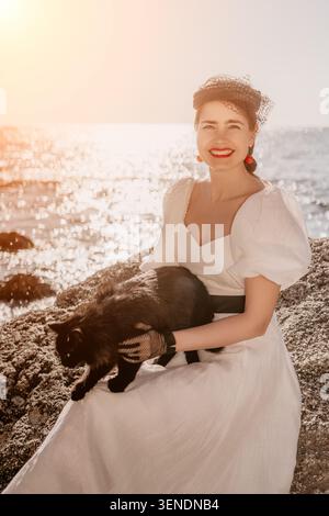 Donna gatto spiaggia sorridente e seduta su rocce con abito bianco e velo al tramonto in riva al mare Foto Stock