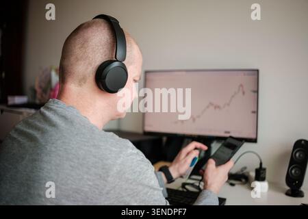 Uomo che indossa le cuffie che utilizza smartphone e computer per il trading finanziario Foto Stock