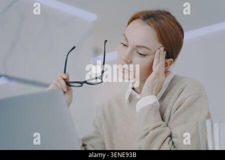 Una donna d'affari stanca tiene gli occhiali mentre lavora con un computer portatile in un ufficio moderno Foto Stock