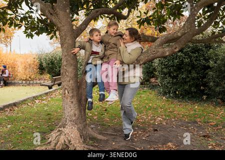 Madre che sostiene i bambini mentre salgono insieme su un albero nel parco cittadino autunnale Foto Stock